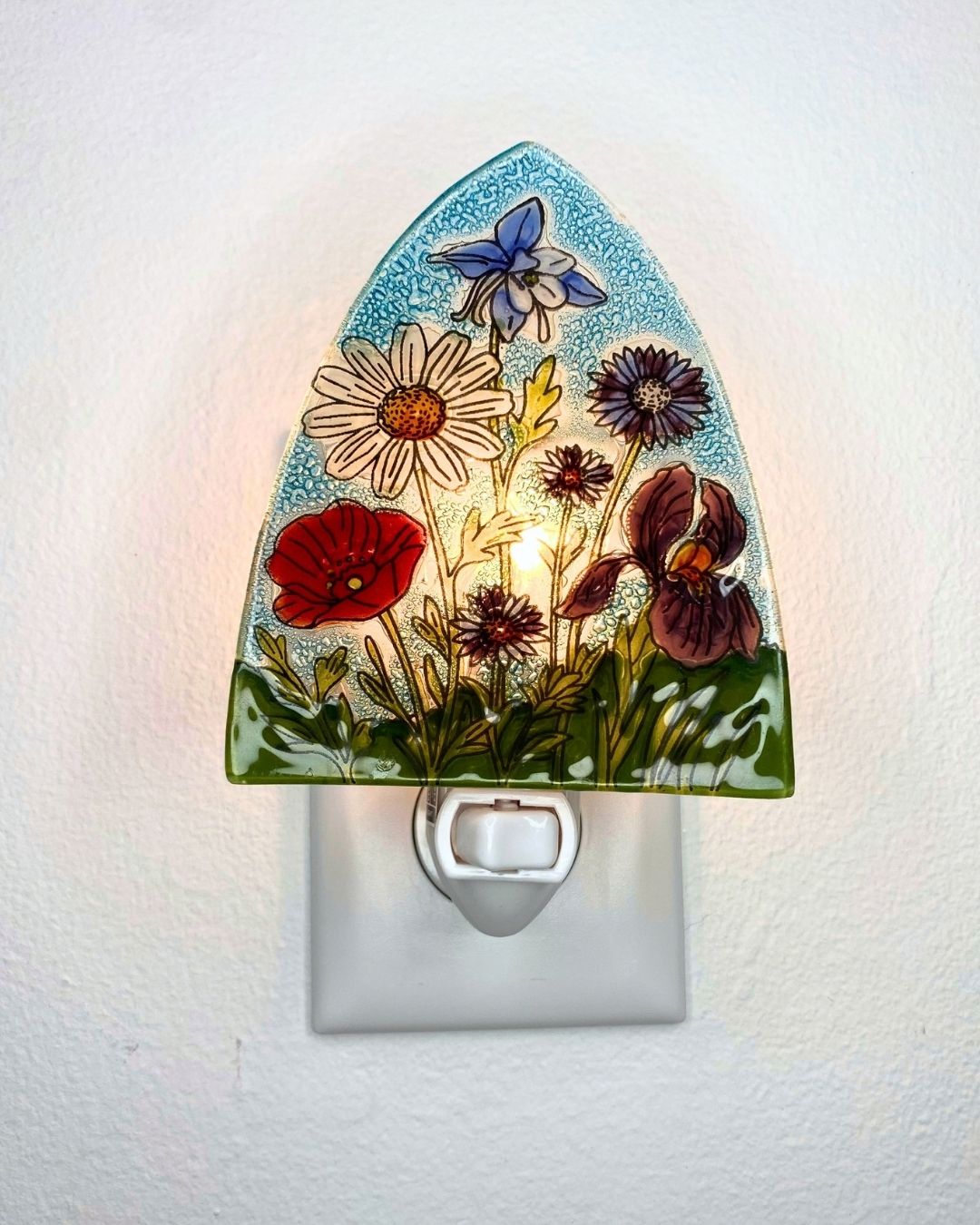 A lit up decorative glass night light with a hand-painted floral design featuring a blue columbine, white daisy, red poppy, and purple pansy on a light blue speckled background, shown plugged into a white wall outlet.