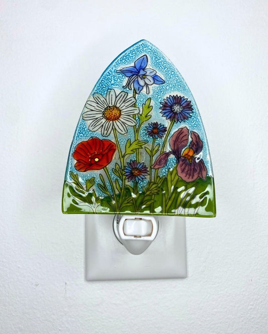 A decorative glass night light with a hand-painted floral design featuring a blue columbine, white daisy, red poppy, and purple pansy on a light blue speckled background, shown plugged into a white wall outlet.