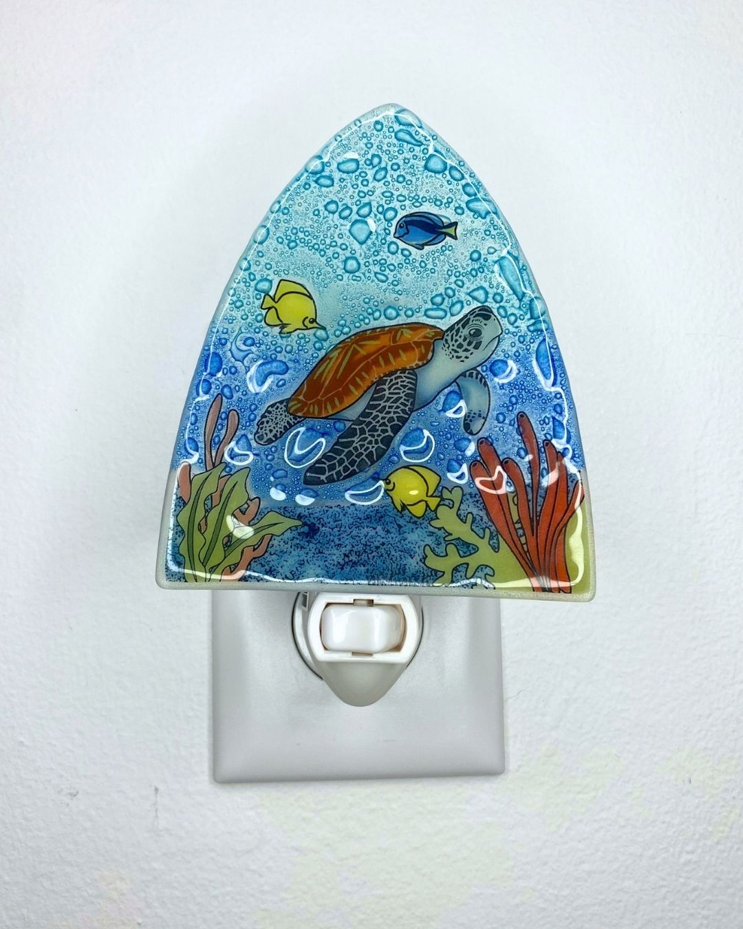 A triangle-shaped glass night light featuring a colorful underwater scene with a sea turtle, yellow fish, and coral on a blue background, shown plugged into a white wall outlet.