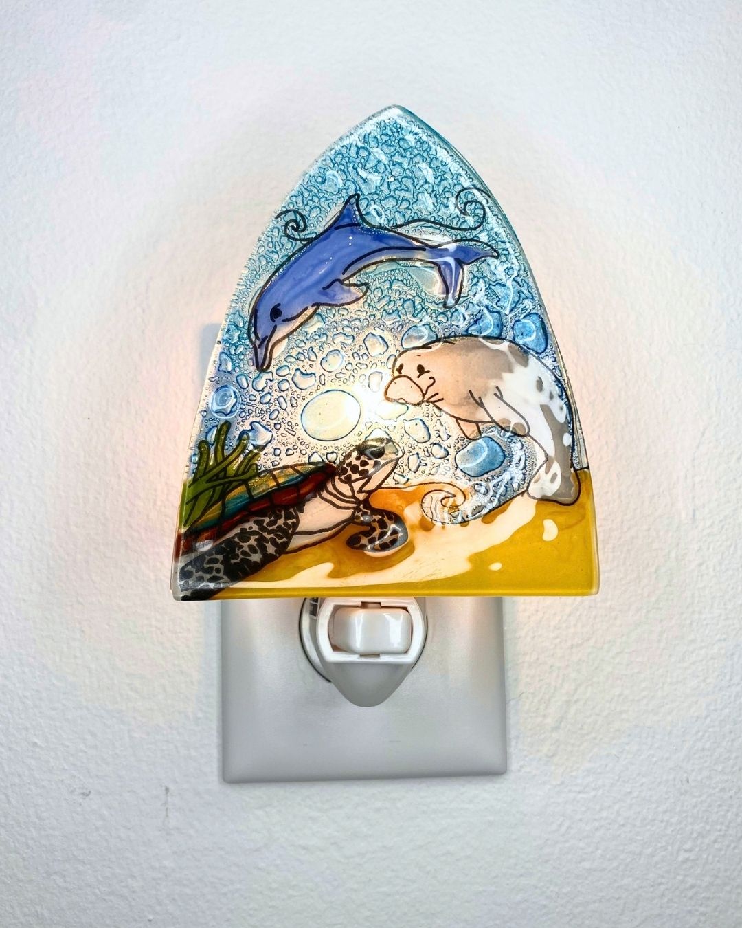 A lit up triangular shaped nightlight featuring a blue dolphin, grey manatee and sea turtle against blue waves and a tan sandy ocean floor.