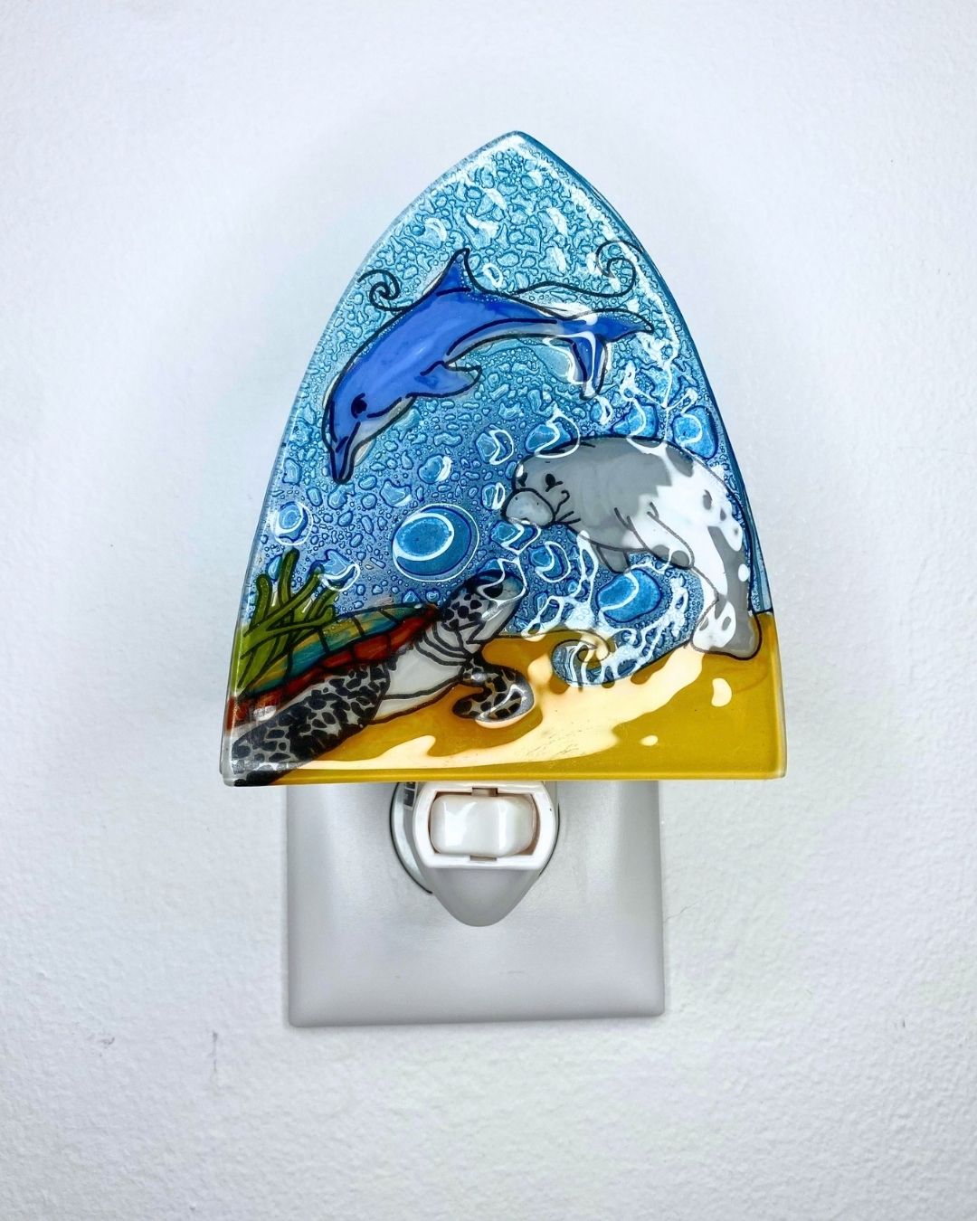 A decorative triangular night light plug-in featuring a blue dolphin, grey manatee, and sea turtle set against ocean waves, bubbles, and a sandy seabed, mounted on a white wall.