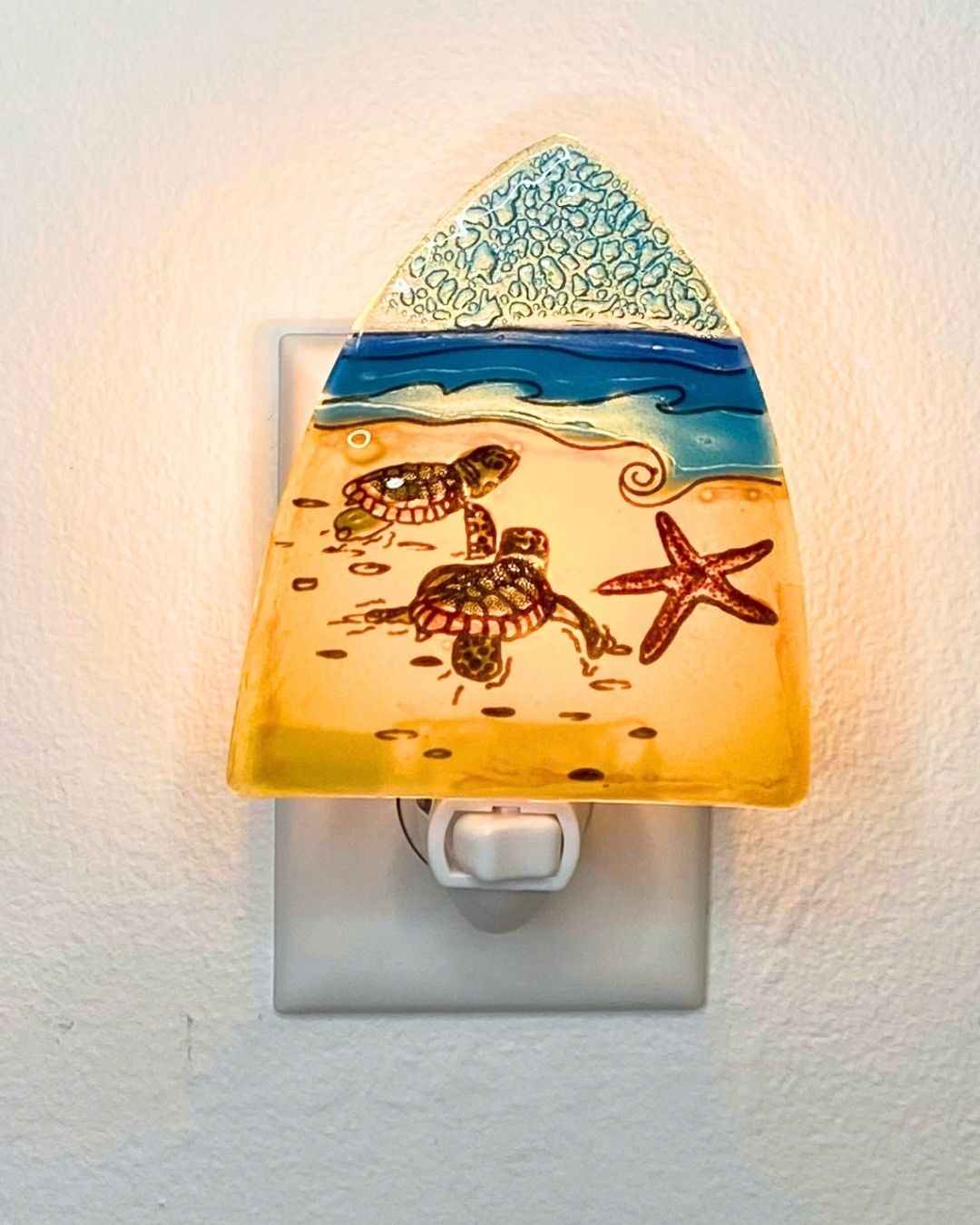 A beach-themed glass night light plug-in featuring sea turtles, a starfish, and ocean waves under a textured sky, glowing warmly on a white wall.