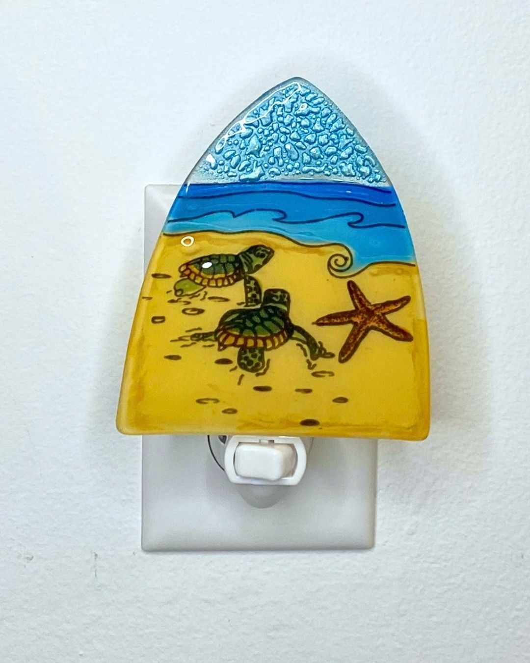 An unlit triangular shaped nightlight is plugged into a white wall. The nightlight features two green sea turtles on yellow sand crawling to blue waves in the distance.  A pink starfish rests on the sand nearby.