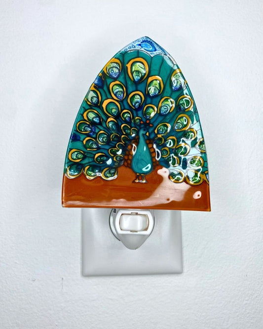 A decorative glass night light shaped like a peacock with a teal and green feathered tail, gold eye details, and a reddish-brown base, shown plugged into a white wall outlet against a white wall.