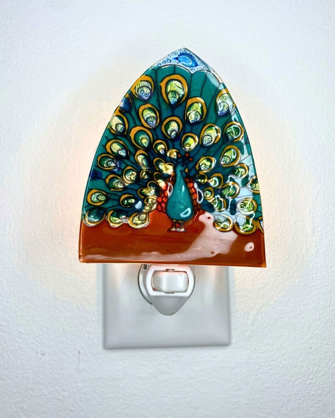A lit decorative glass night light shaped like a peacock with a teal and green feathered tail, gold eye details, and a reddish-brown base, shown plugged into a white wall outlet against a white wall.