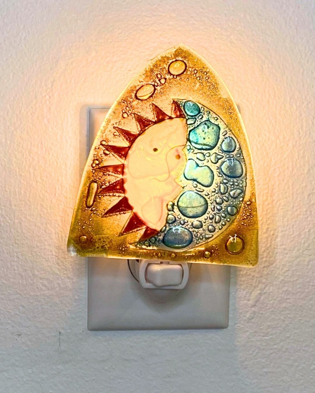 A decorative fused glass night light shaped like a crescent moon and sun, glowing warmly against a white wall. The design features a light blue moon and a yellow sun on a golden background.