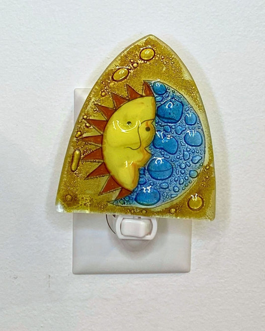 A triangular shaped nightlight is plugged into a white wall. The design features a half-sun, half-moon design on a golden background.