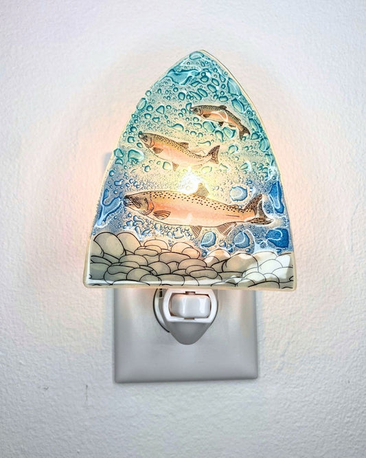 A fish-themed night light mounted on a white wall, featuring three salmon swimming through blue water with rocks at the bottom, shown illuminated from within.