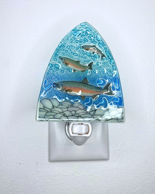 A decorative triangular glass night light, featuring three trout swimming in blue water with rocks at the bottom, shown plugged into a white wall outlet.