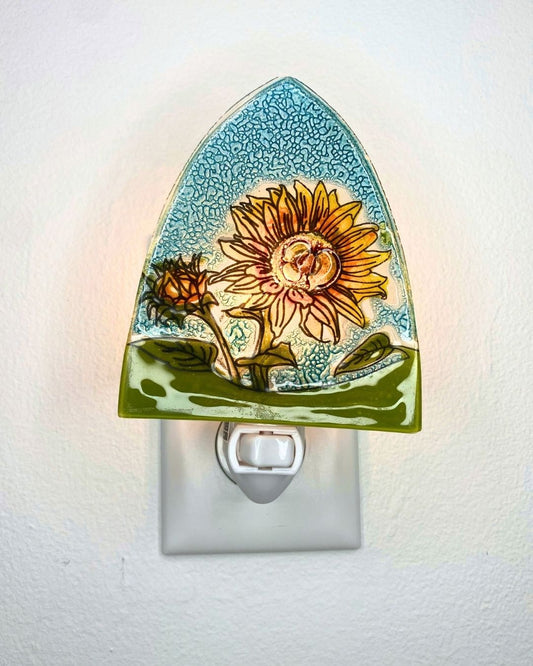 A decorative glass night light with a sunflower design, shown plugged into a white wall outlet. The glass features a glowing sunflower with yellow petals, green leaves, and a textured light blue background.