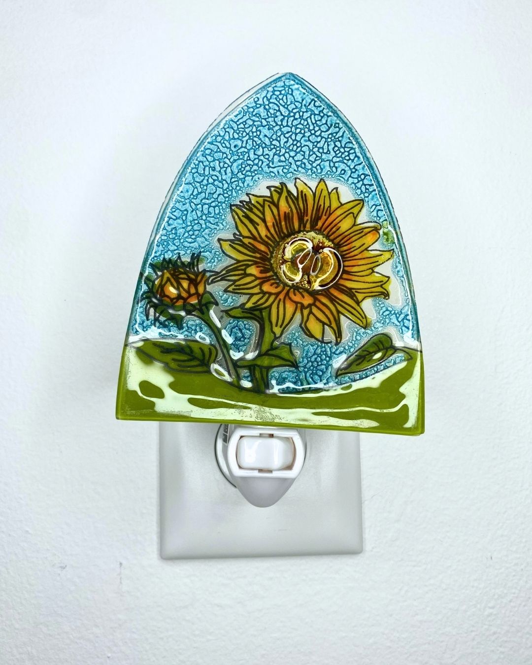 A decorative glass night light plug-in featuring a painted sunflower and green leaves on a textured blue glass background, shown mounted on a white wall outlet.