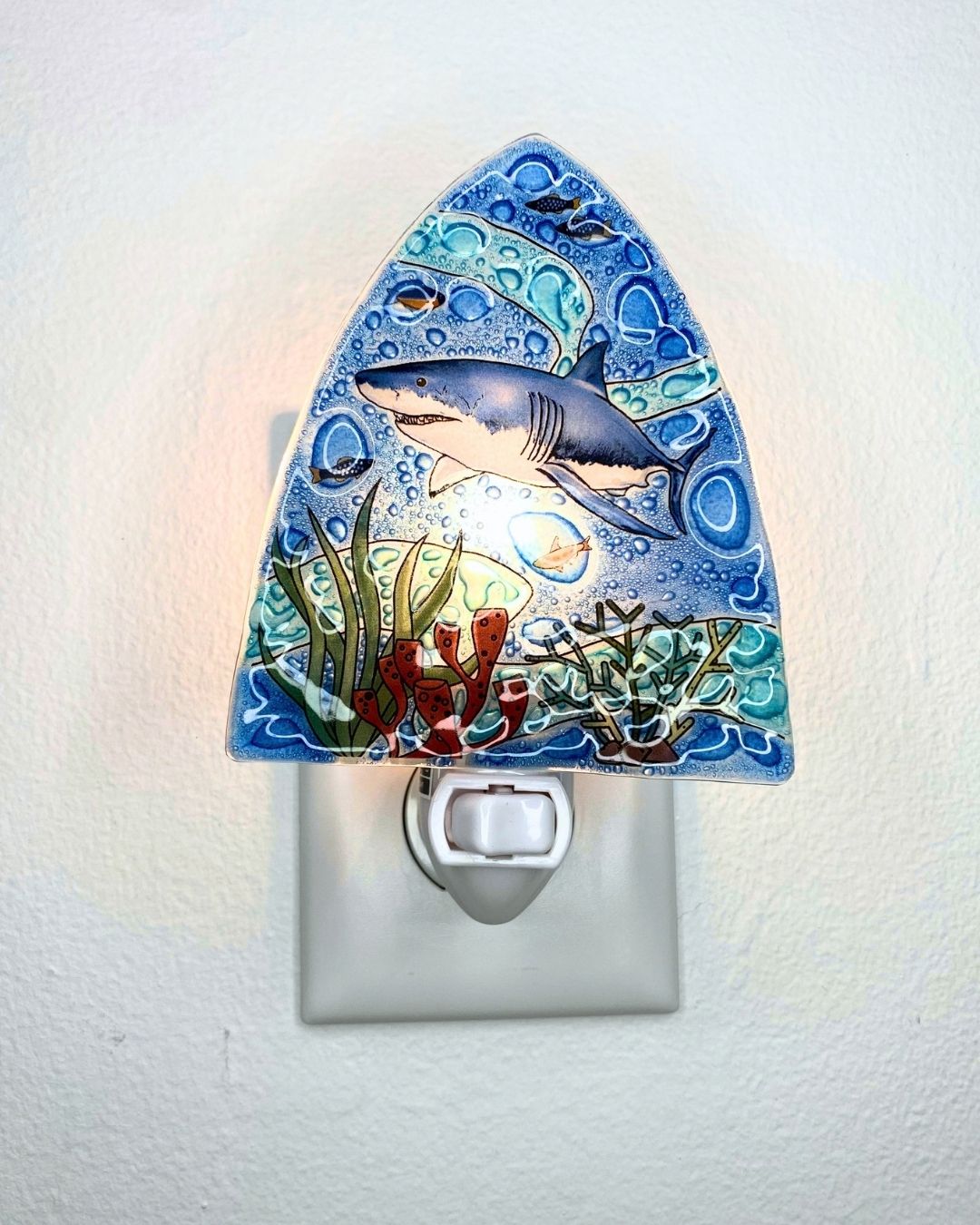 A decorative night light shaped triangle, mounted on a white wall plug against a white textured wall. The night light features a blue and gray shark, green seaweed, and red coral details, with a warm glow emanating from within.