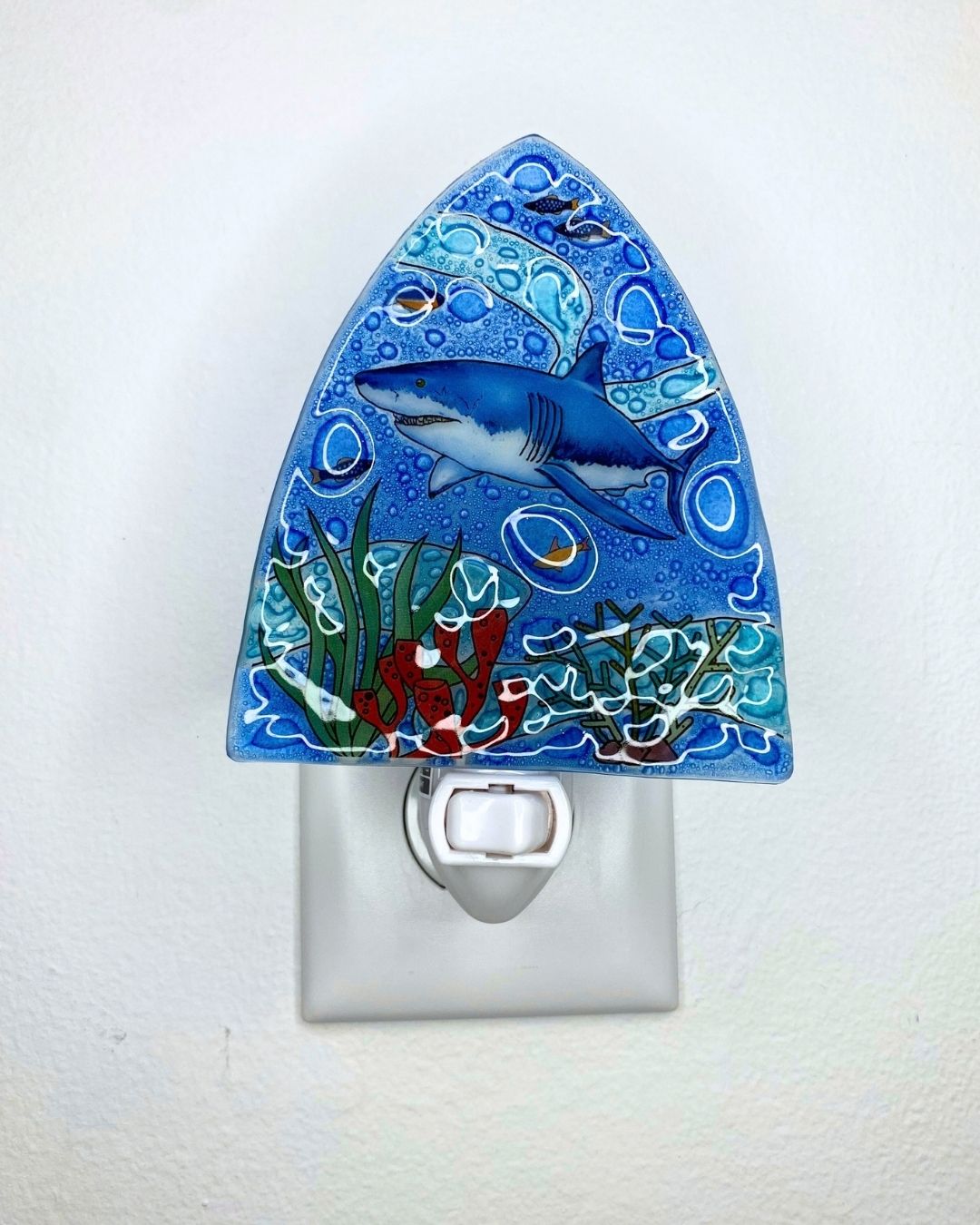 A decorative blue night light plug-in with a hand-painted shark and underwater scene, shown mounted on a white wall.