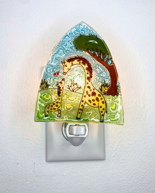 A decorative glass night light lit up and featuring a giraffe and tree design, plugged into a white wall outlet against a white textured wall.