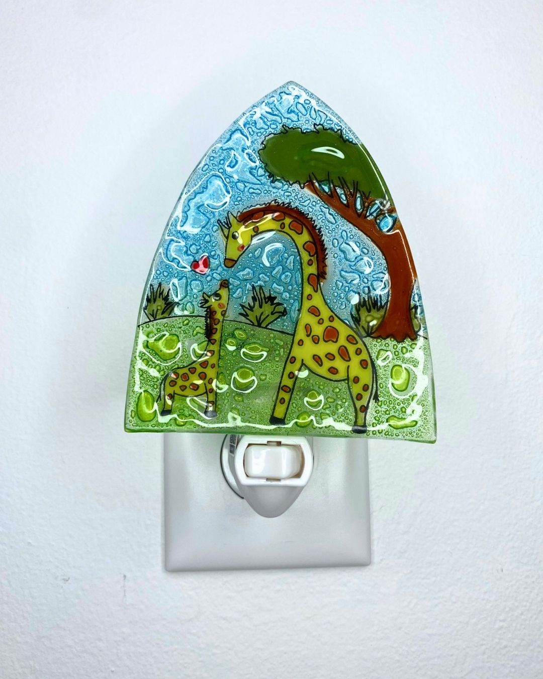 A decorative glass night light with an African safari design featuring a mother and baby giraffe, a tree, and a blue sky, shown plugged into a white wall outlet.