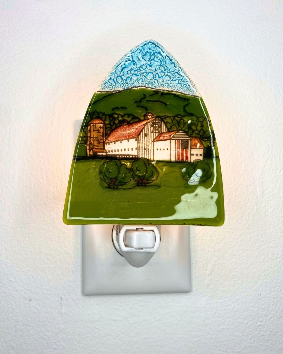 A lit triangular nightlight featuring a white barn against a green hill and flying the American flag is plugged into a white wall.