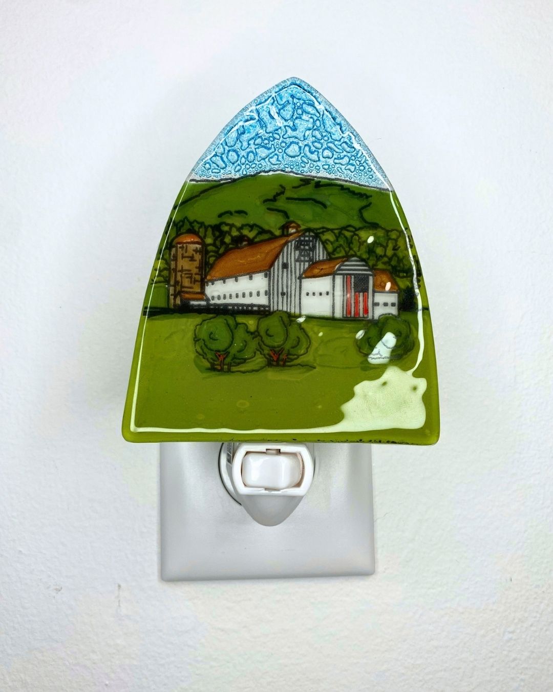 An unlit triangular nightlight featuring a white barn against a green hill and flying the American flag is plugged into a white wall.