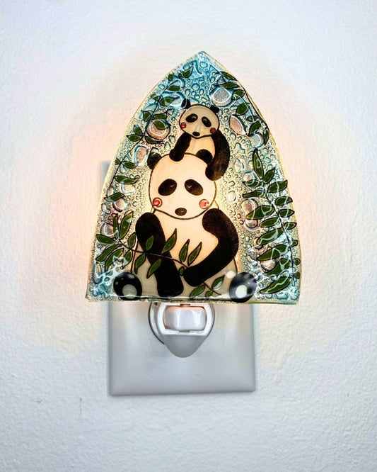 A lit up decorative glass night light featuring a panda mother and panda baby design with bamboo leaves, shown plugged into a white wall outlet against a white wall.
