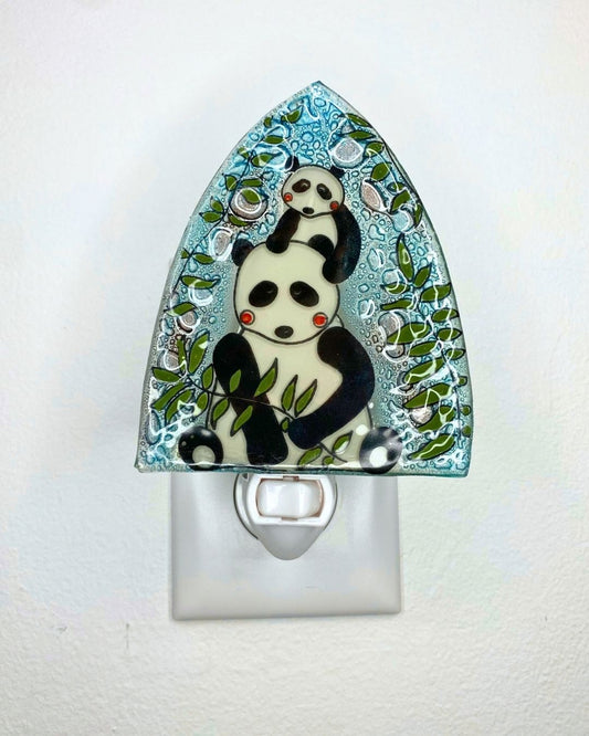A glass night light with a decorative arched top featuring a mother and baby panda illustrations and green bamboo on a light blue patterned background, shown plugged into a white wall outlet.
