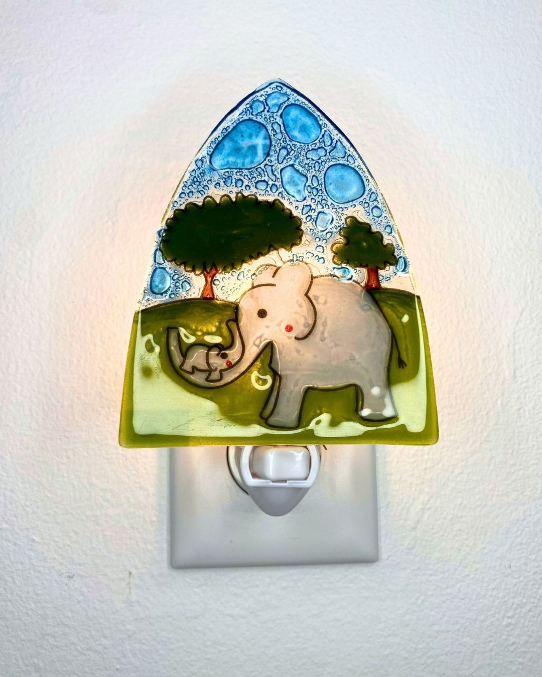 A decorative glass night light shaped like a triangle, featuring a gray mother elephant and baby elephant on a green landscape with blue patterned sky, shown plugged into a white wall outlet and glowing from inside.