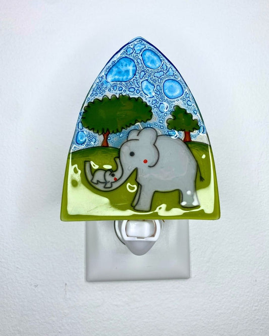 A decorative glass night light plug-in featuring a painted scene of a gray mother elephant and baby elephant under green trees, with blue patterned sky and a light green base, shown plugged into a white wall outlet.