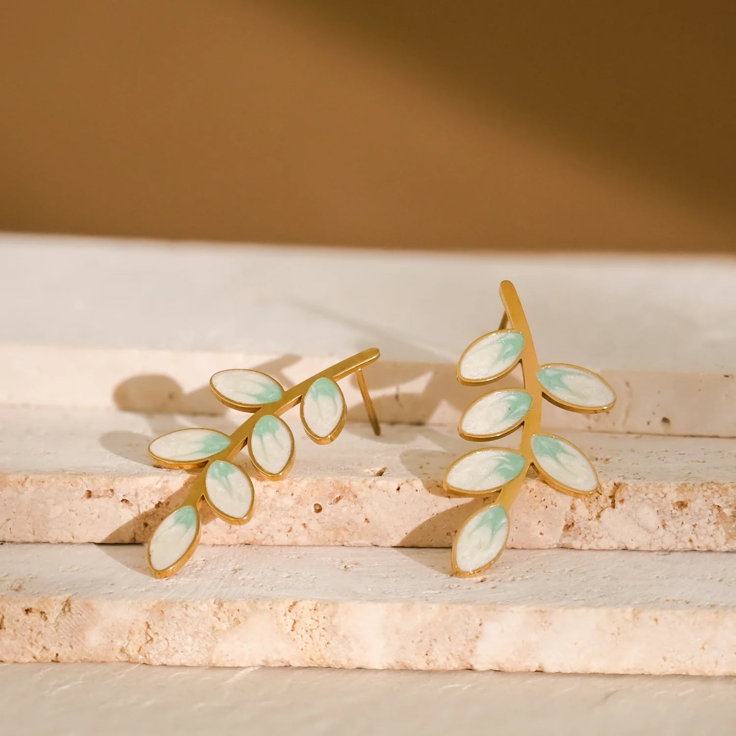 Gold leaf-shaped earrings with green accents styled on a tan stone surface.