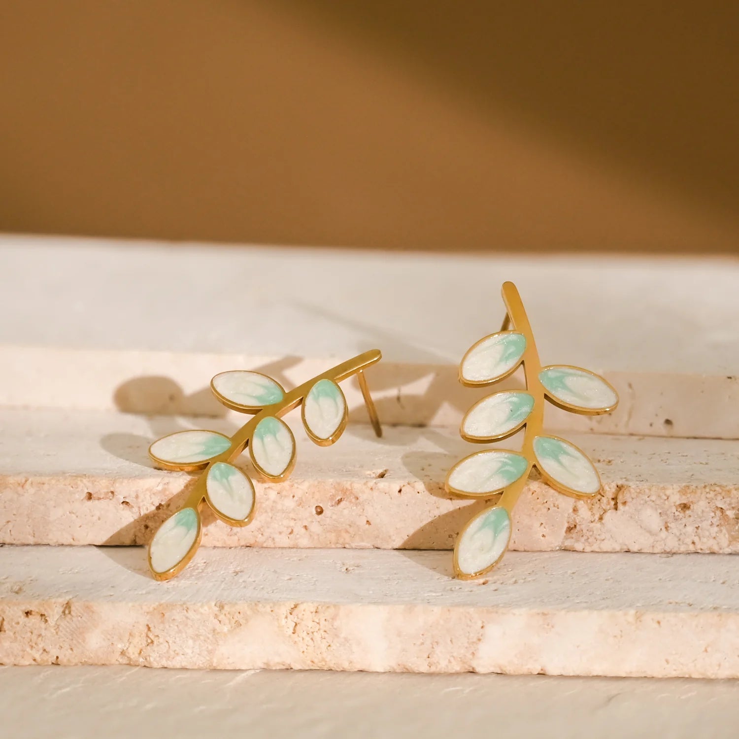 Gold leaf-shaped earrings with green accents styled on a tan stone surface.