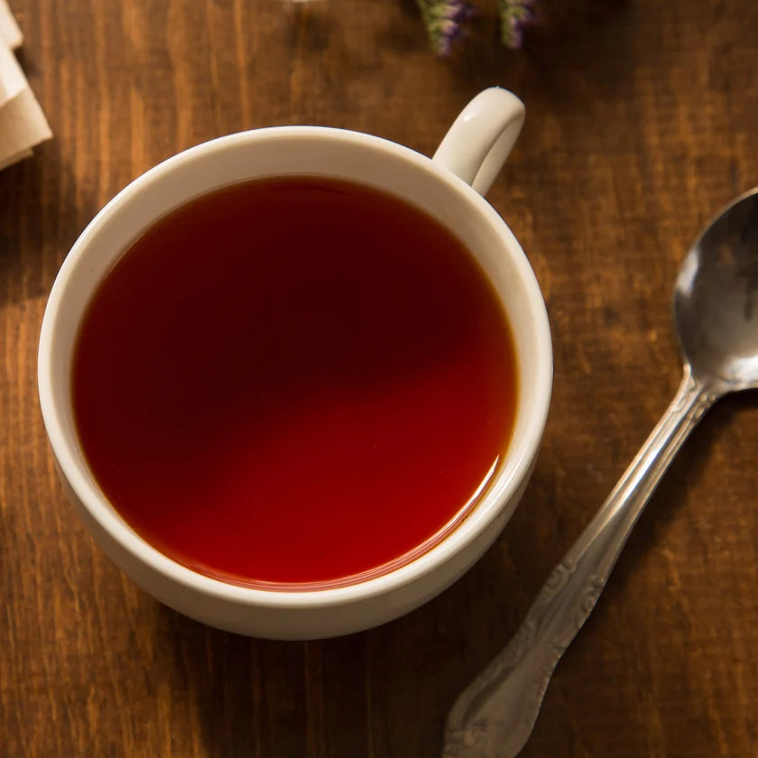 A white ceramic cup filled with a deep red tea, placed on a dark wooden table next to a silver spoon.