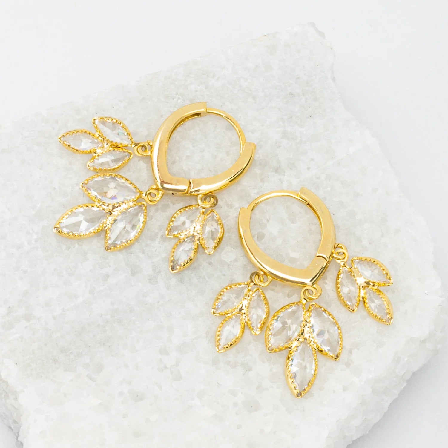 Gold hoop earrings with leaf-like sparkly drops on a white textured background