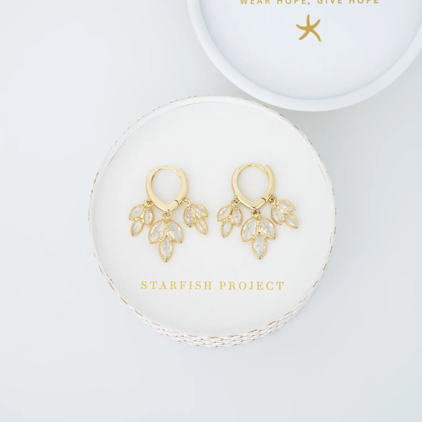 Gold huggie earrings with clear teardrop dangles in a Starfish Project branded box with a white background
