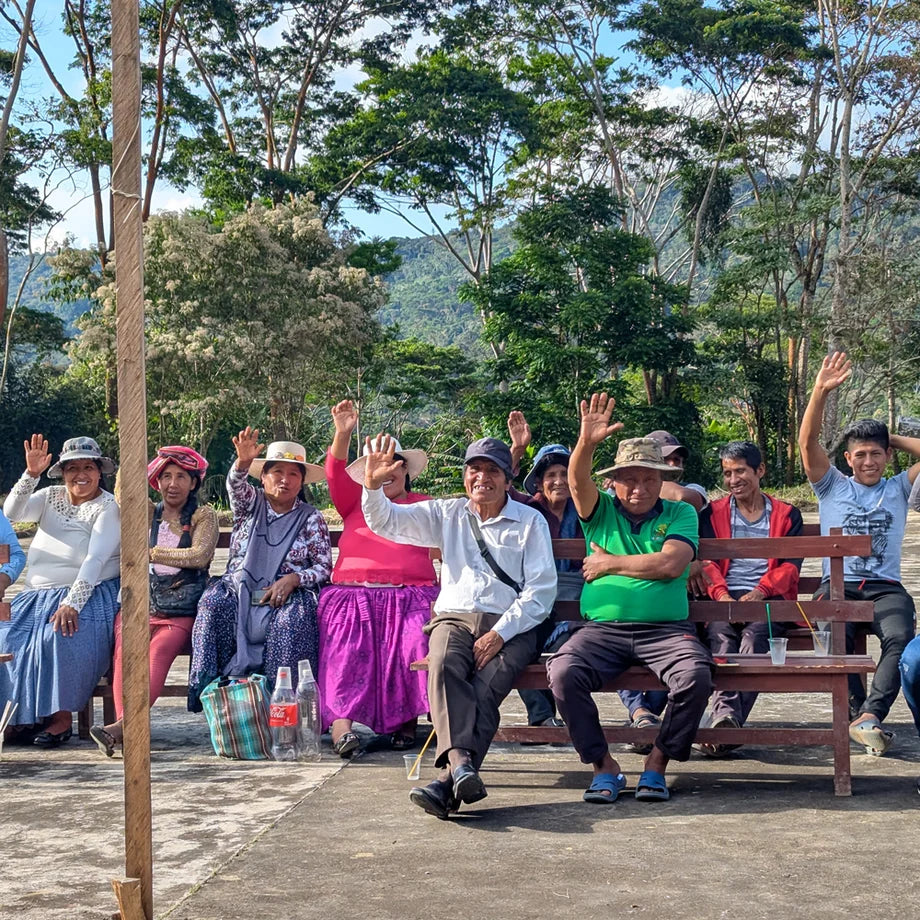 Ten Bolivian farmers are shown raising their hands to wave at the camera while sitting on benches outdoors.