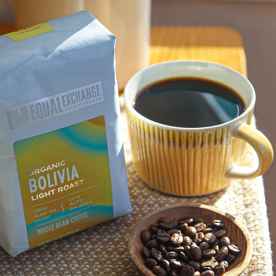 A white and yellow bag of coffee sits on a table next to a small bowl of whole coffee beans and a yellow cup full of coffee.