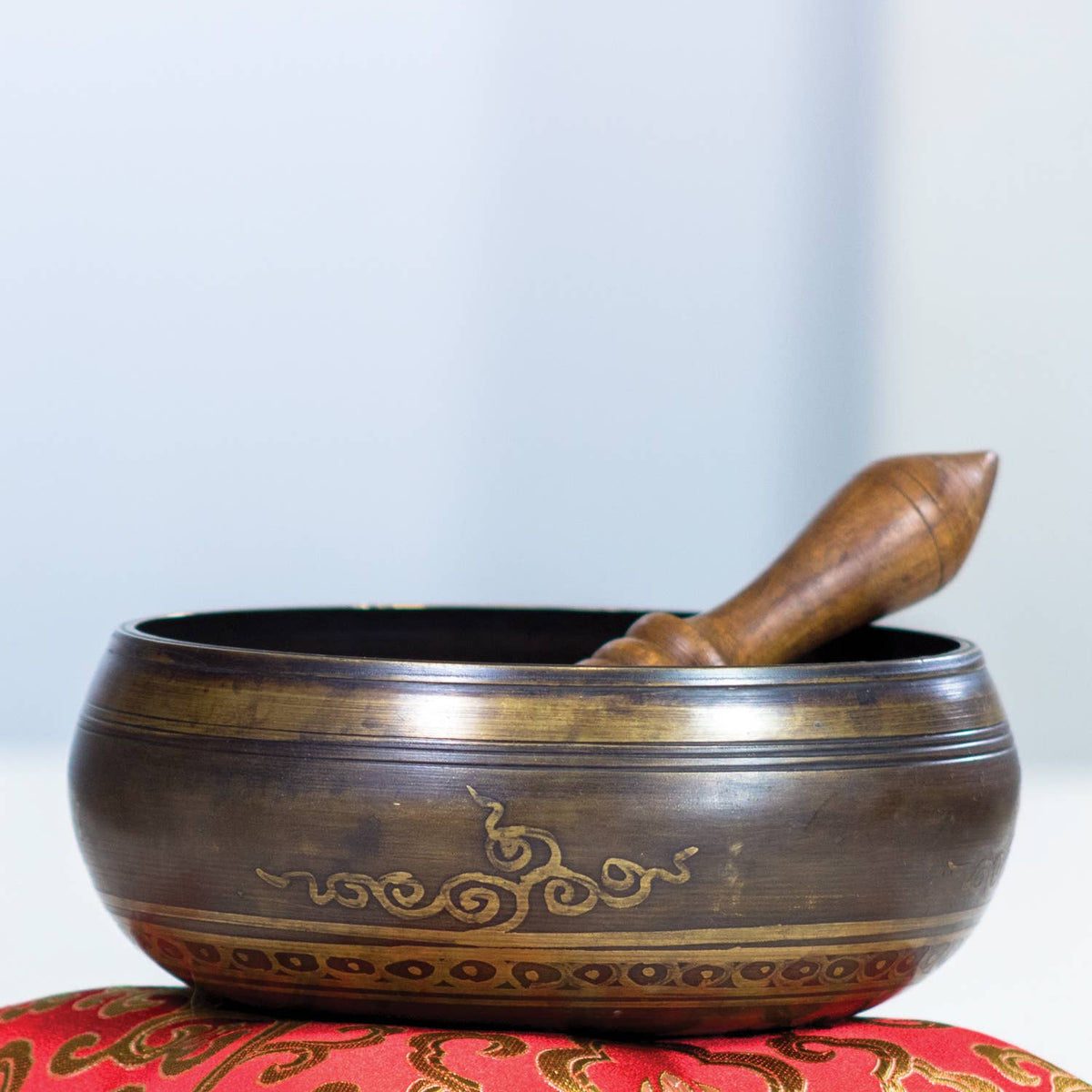 Tibetan singing bowl with wooden striker on a red cushion against a light blue background