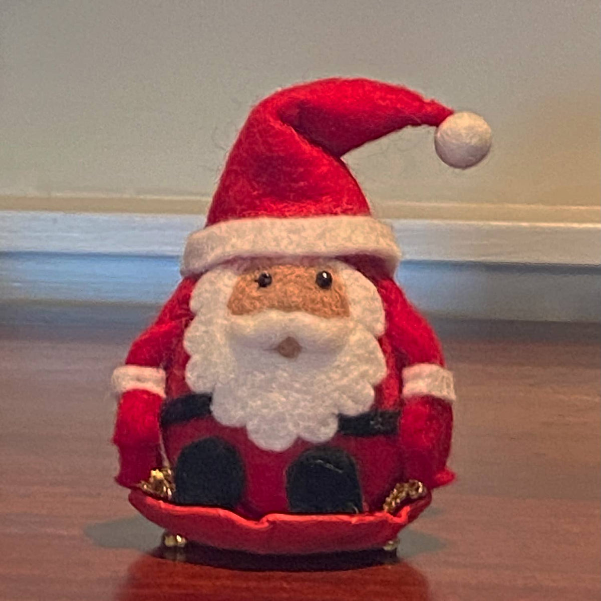 A small felt Santa Claus decoration sitting on a wooden surface against a plain indoor wall. The Santa figure is red with a white beard, white trim, black boots, and gold accents.