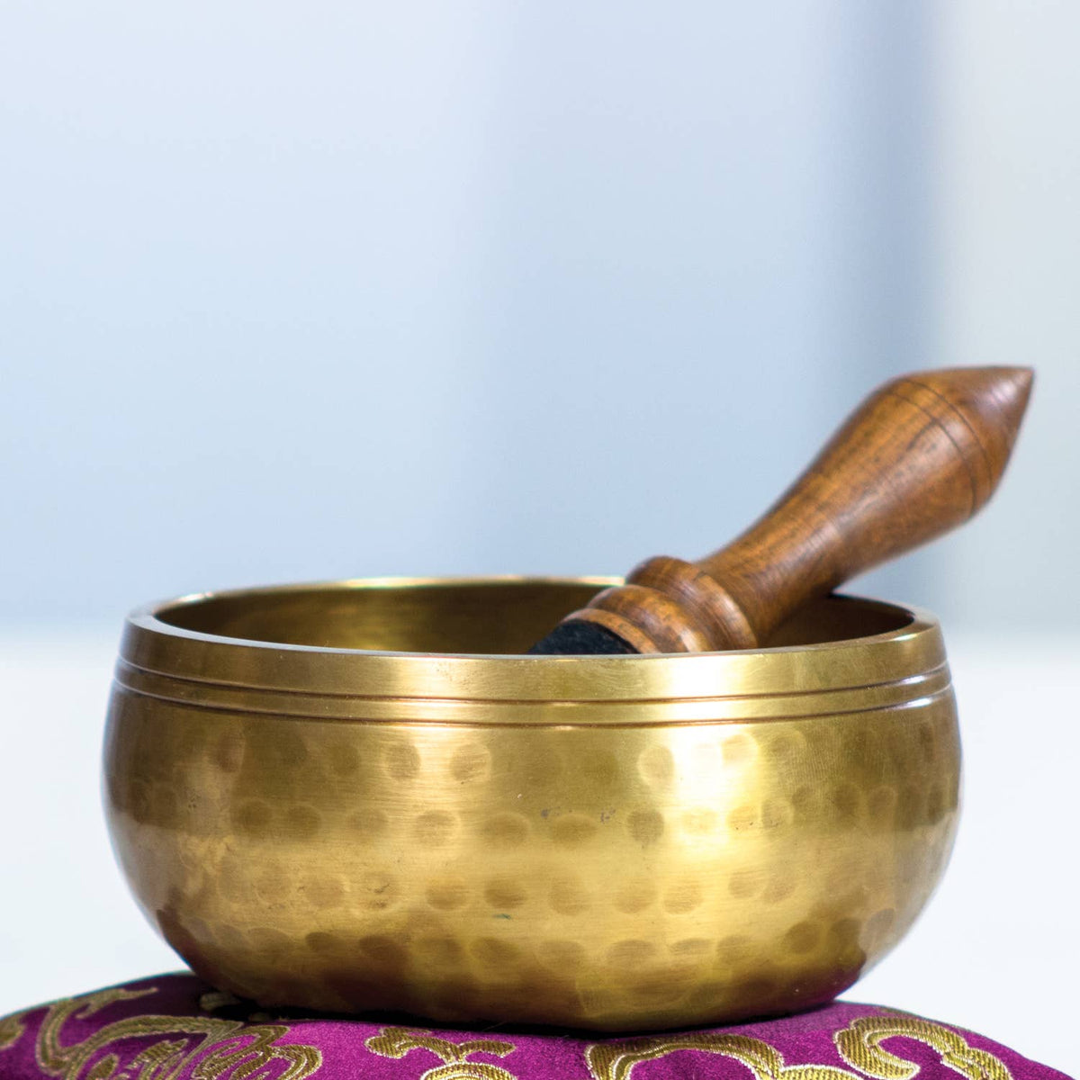 Brass singing bowl with wooden striker on a purple cushion against a light blue background