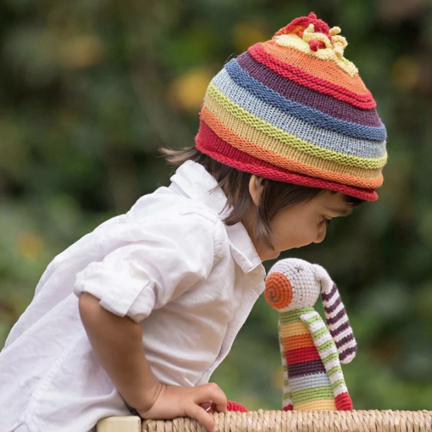 A child with a rainbow crochet hat with a similarly colored stuffed rabbit outdoors.