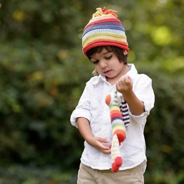 A child with a rainbow hat plays with a rainbow stuffed rabbit outdoors.