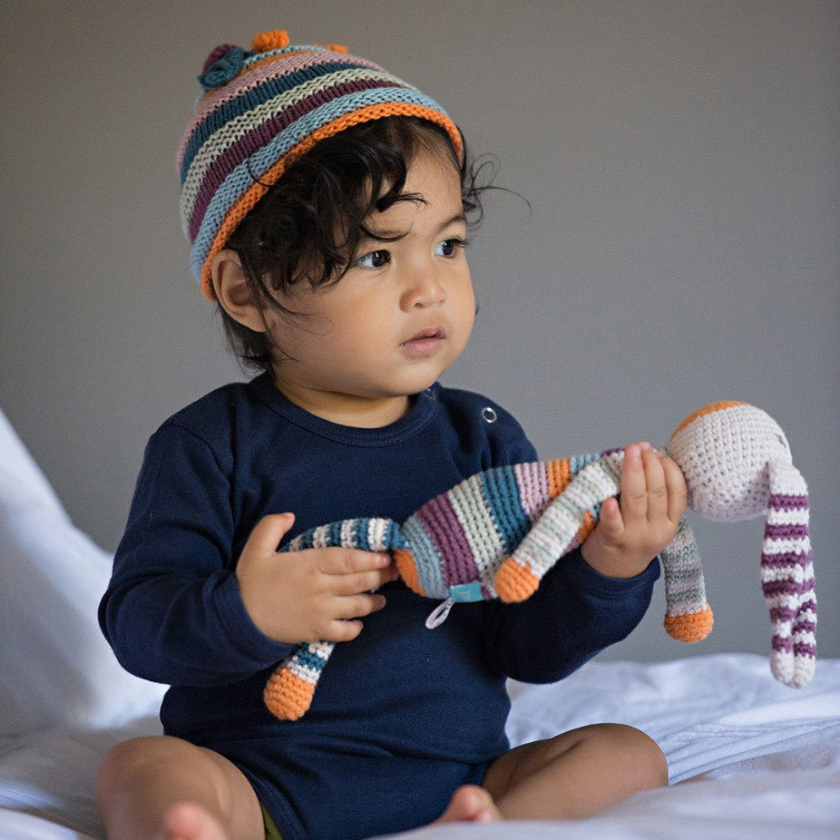 Child wearing a colorful knit hat holding a striped toy on a bed.