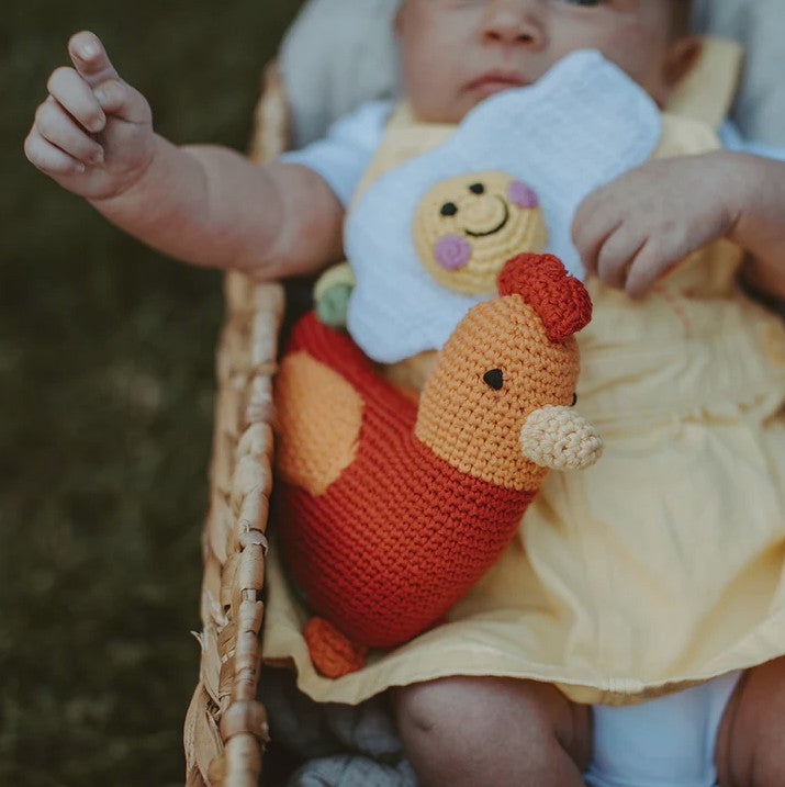 Baby in a yellow dress holding a knitted chicken toy with a smiley face bib.