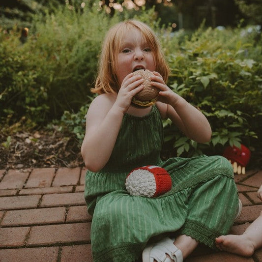A red-head toddler wearing a green dress is shown taking a big "play" bite of a crocheted burger toy.