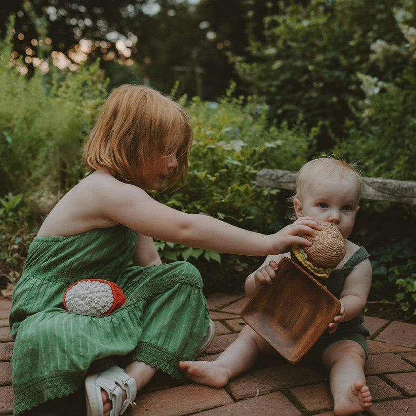 Two children playing outdoors on a brick path with greenery in the background. One "feeds" the other a crocheted burger toy to eat.