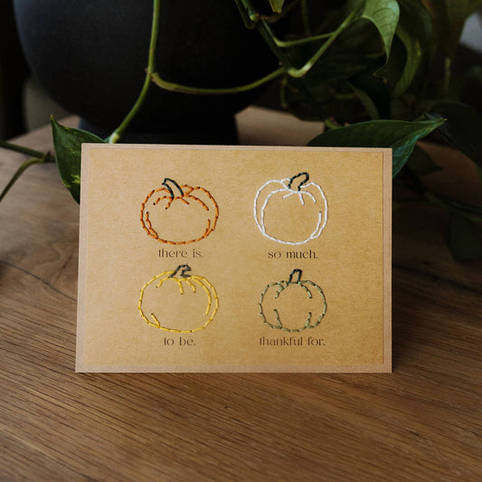 A square kraft paper card with four embroidered pumpkin illustrations and the phrases 'there is', 'so much', 'to be', and 'thankful for.' in black text, displayed on a wooden surface with green plant leaves in the background.