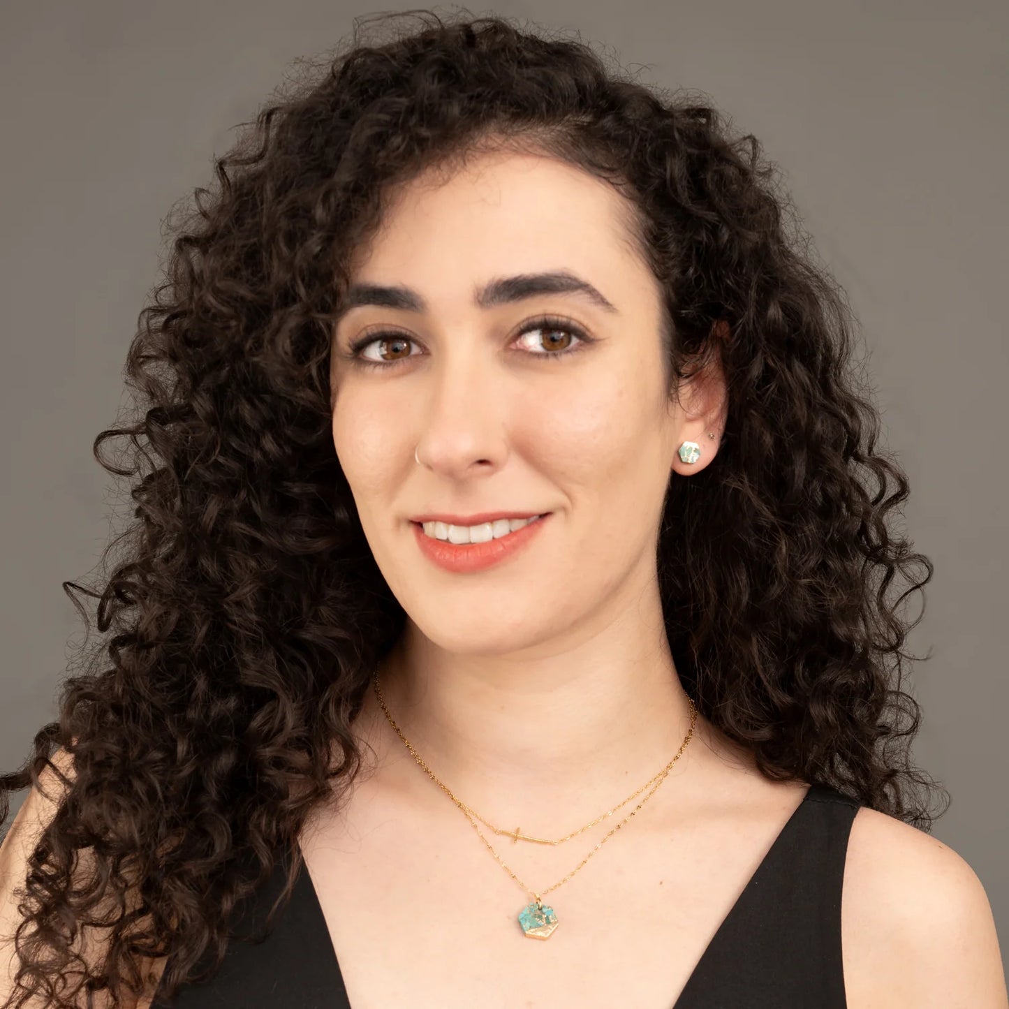 Woman with curly hair wearing a black top and two gold necklaces against a gray background