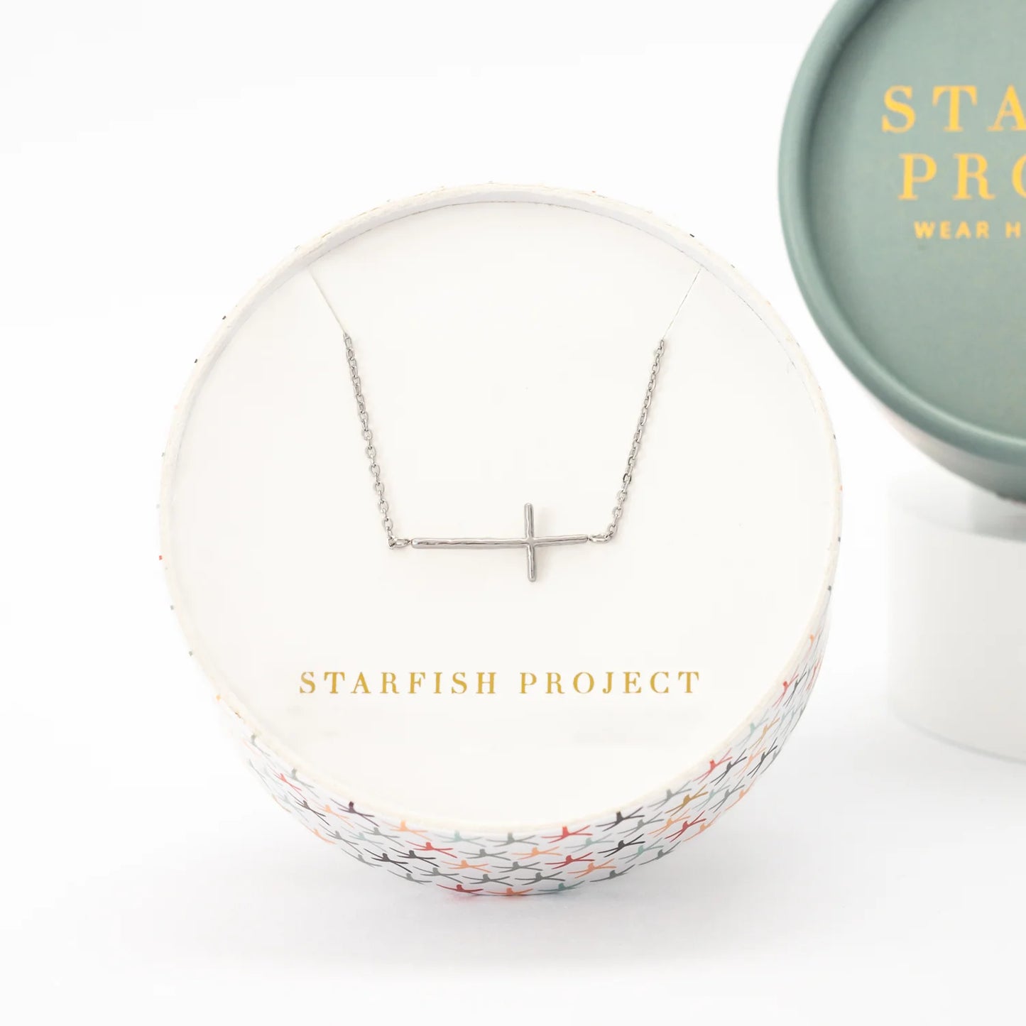 A silver necklace with a minimalist horizonal cross pendant displayed inside a round white gift box. The box features gold lettering that reads 'STARFISH PROJECT'.