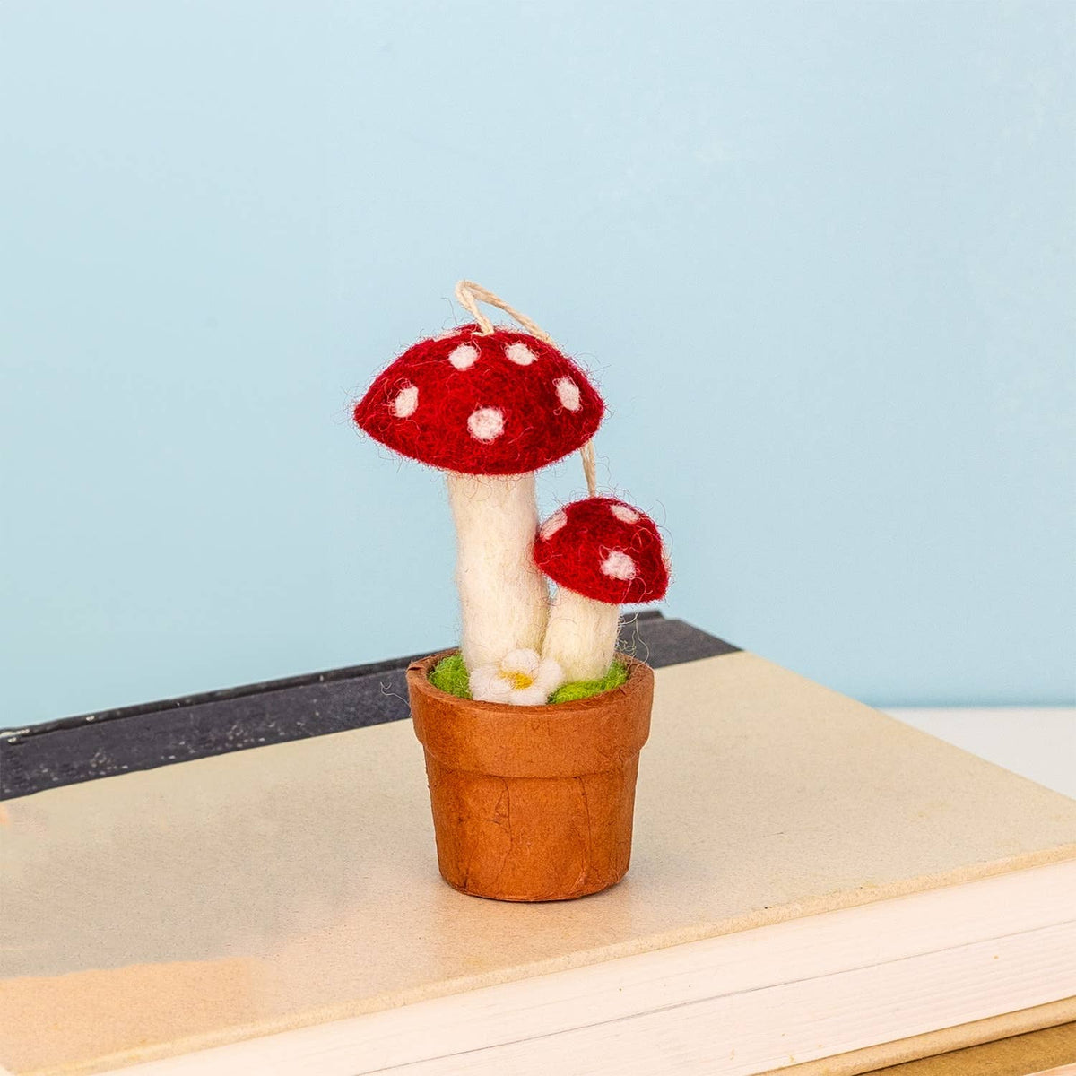 A miniature felted wool mushroom decoration in a small brown pot, displayed on top of a book against a light blue background.