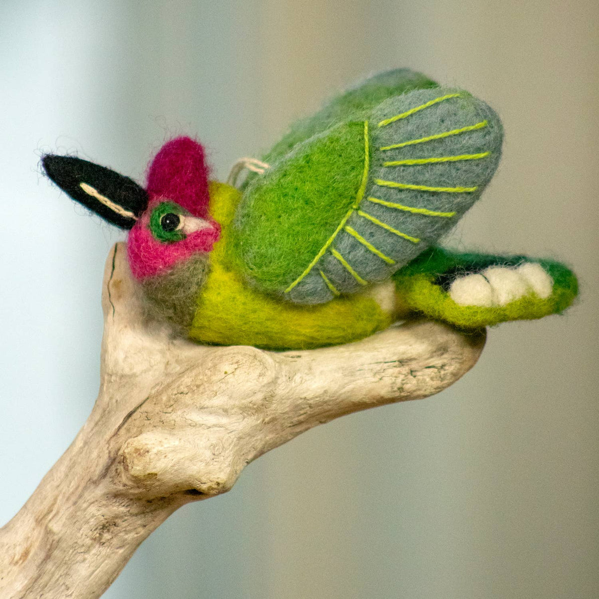 A handcrafted felted wool bird ornament perched on a natural driftwood branch. The bird features a bright lime green body, gray wings with yellow-green stitched details, a magenta-pink beak, and a black-and-white striped crest.