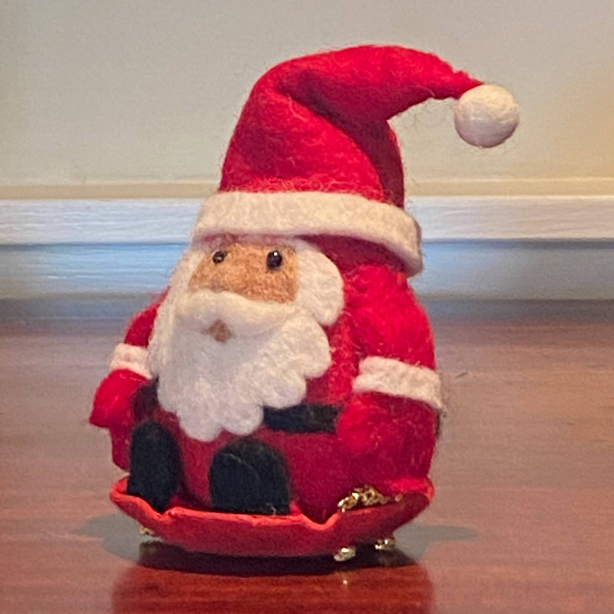 A red and white felt Christmas Santa Claus figurine sitting in a red sleigh on a wooden surface.
