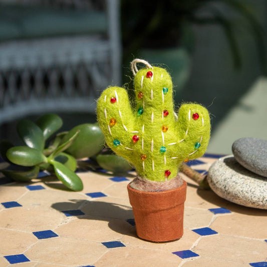 A felted green cactus ornament in a small terracotta pot, decorated with colorful rhinestones and white stitching, set on a tiled surface outdoors.