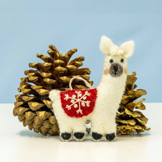 A white felt llama ornament with a red belly patch featuring a white snowflake design, posed between two gold pinecones against a light blue background.