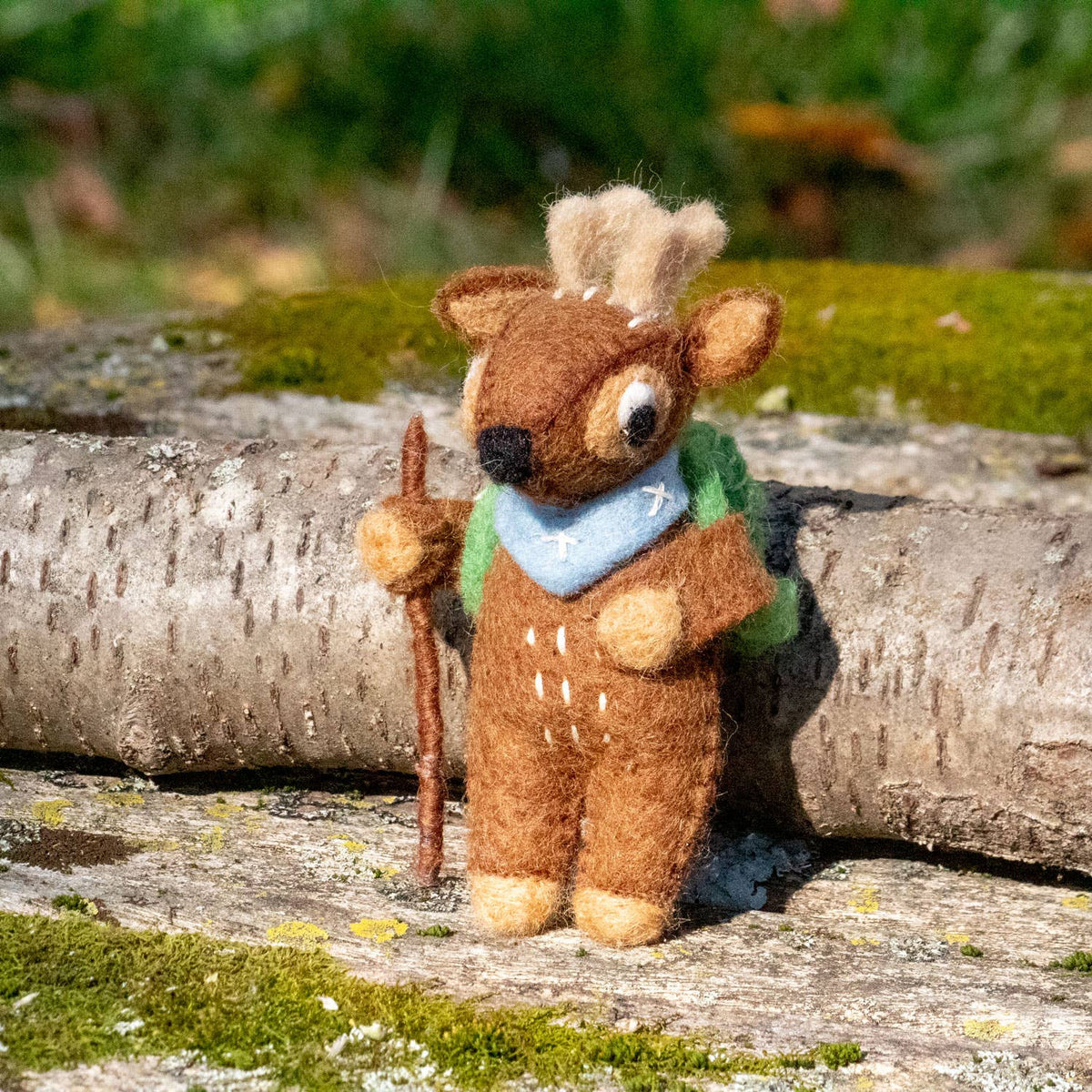 Stuffed deer ornament with a green backpack and walking stick on a wooden log with a natural background